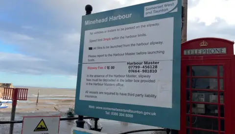 A board at the harbour lists various details. It states 'no vehicles or trailers to be parked on the slipway', 'speed limit 3mph within the harbour limits', jet skis to be launched from harbour slipway' and 'please report to the harbour master before launching boats'. Below that is the fee information and the contact information. Next to the sign is a red phone box, and to the left is the harbour at low tide and the Bristol Channel. 