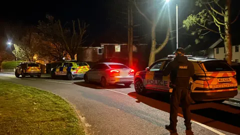 A road with three police cars on it. It is dark and houses are in the background. A PSNI officer is standing with his back to the camera.