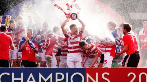 A man in a red and white football kit lifts a trophy, surrounded by other players from his team. Several people are spraying bottles of champagne. A sign reads Champions 2025 in the foreground.