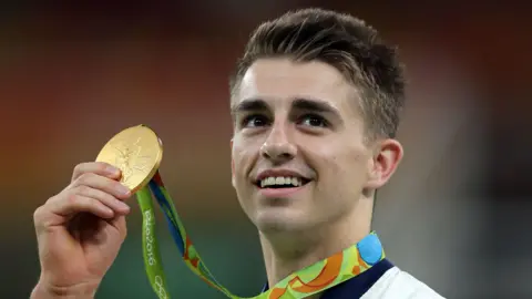PA Media Max Whitlock holds an Olympic gold medal in the air at the 2016 games in Rio. It is attached to a green, orange and blue ribbon. He has short brown hair and is smiling.