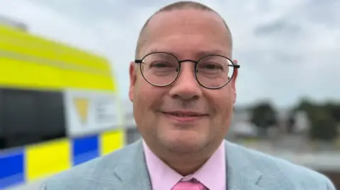 A man wearing round glasses, a grey suit and pink shirt and tie. He is standing outdoors in front of a blue and yellow van under a grey sky.