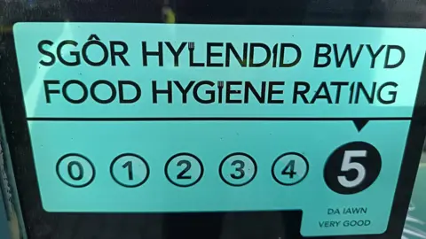 a food rating sign in a glass window with the words in black on a green backghroun saying Food hygiene rating in welsh and english abover the numbers 0 to five witth circles around them and the five in a black circle wih and arrow above and very good in Welsh and english 
