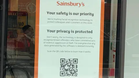 Warren Rajah A Sainsbury's poster in a store's front window, outlining its use of facial recognition technology, above a QR code.