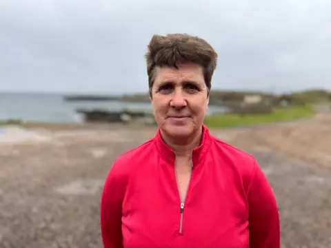 A woman with short brown hair in a pink jumper stands in front of a blurred background that includes green grass, a stoney foreground and sea in the background.