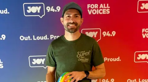Joy 94.9 A man wearing a cap and t-shirt and holding a rainbow football smiling at camera in front of a backdrop with Joy 94.9 radio station branding. 