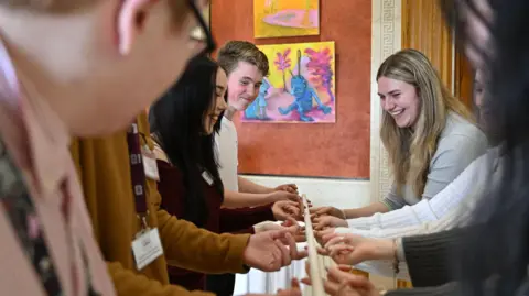 Michael Cooper Young people are lined up holding a stick together. A blonde girl smiles on one side of the picture while a brown haired girl and brown haired boy smile fm the other side of the picture. 