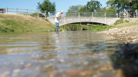 Jamie Niblock/BBC A man stands in a river with the water up to his thighs. He is wearing wellington boots. The water stretches out from the camera and a bridge is in the background.