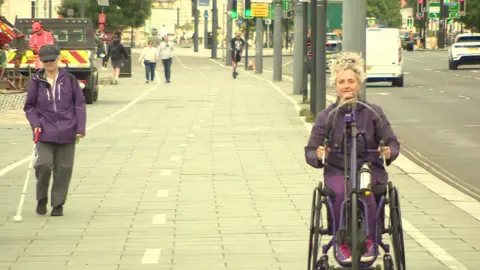 BBC Terri Ballon walks along the Strand using a white cane to guide her. On the right Dr Kay Hinkle rides her arm-pedal bike 