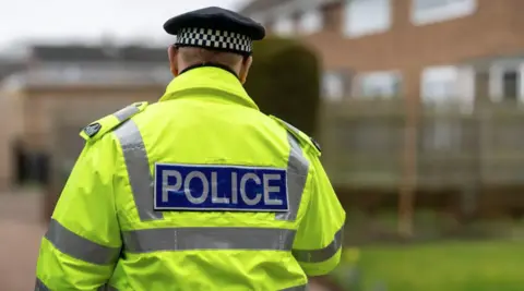 The back of a police officer in a yellow high visibility coat. There are buildings and a lawned area in front of him, blurred.