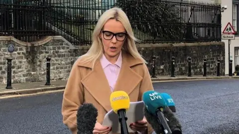 PA Media DCI Duffie reading as she stands in front of four different mics. She has long blonde hair and is wearing black glasses, a pink shirt and a brown coat.