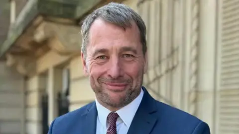 Councillor Stephen Atkinson standing outside County Hall in Preston smiling. He has short fair hair and stubble and is wearing a red rose badge on his navy blue suit jacket.
