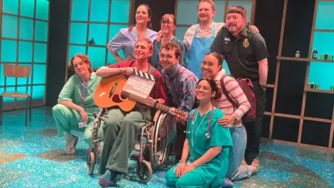 A group of nine performers in medical scrubs and various outfits pose together on the teal-patterned stage floor. One performer in a wheelchair holds an acoustic guitar and a clapperboard.