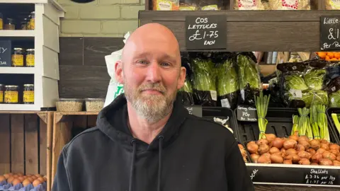 Holly Nichols/BBC Andrew Webb who has a grey and light brown beard and is wearing a black hoody. He is standing in his greengrocers. Behind him are shelves with eggs, honey, lettuces, spring onions and brown onions. 