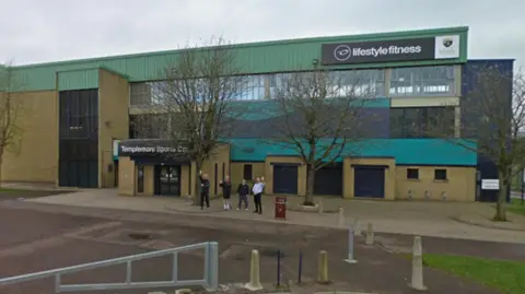Google An outside shot of Templemore Sports Complex in Derry.A large green and brown building. Four people are standing outside waving. 