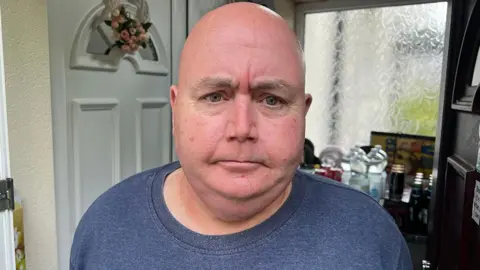 Head and shoulders of Alaster Hirst standing in the doorway of his home. He is bald and is wearing a blue marl top