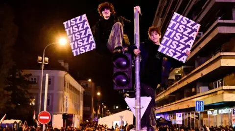 Reuters People hold signs with the name of the opposition Tisza party as others gather after the announcement of partial results of the parliamentary election, in Budapest, Hungary