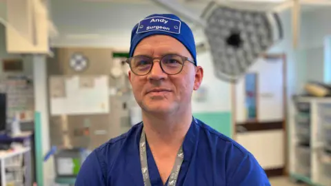 Andy Stevenson looks at the camera as he stands in a clinical room at Musgrove Park Hospital. He is wearing blue hospital scrubs with a blue name badge surgical hat that says 'Andy Surgeon'.