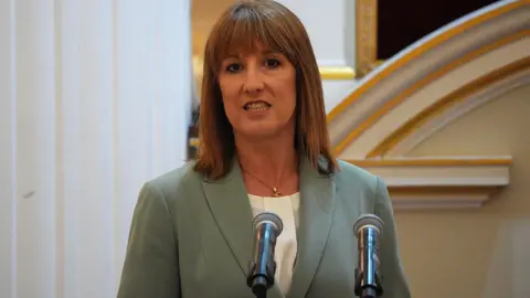 Chancellor Rachel Reeves wearing a sage green jacket and white collarless blouse stands behind a podium with two microphones giving a speech at the annual Mansion House dinner in the City of London, UK. 