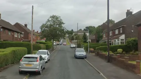 Google Google streetview of houses, gardens and parked cars lining both sides of a single carriageway suburban road.