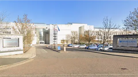 Google A large cream stone building with a glass entrance and large car park. There are two display walls at the entrance to the car park with silver signs reading "Barclays Northampton Campus". A British flag and another blue flag fly from tall poles.