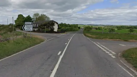 Junction of the A92. There is countryside around the road and a house and sign. 