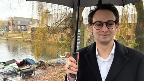 Martin Heath/BBC Josh Dean with medium-length dark hair and black-framed glasses, smiling at the camera while wearing a black jacket and white shirt. He is holding an umbrella. There is a river behind him and the stern of a burnt-out boat is visible behind him. There are houses alongside the river beyond.