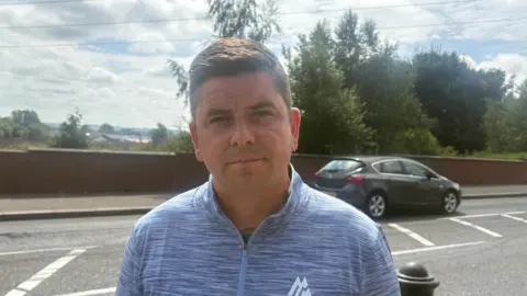 A man with short brown hair looks at the camera. He is standing beside a road and is wearing a light-blue top. There are trees behind him. On the road, a car is passing.