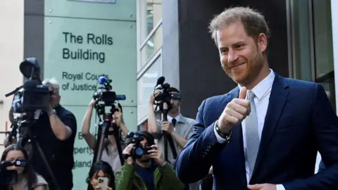 Reuters Prince Harry, wearing a blue suit, blue shirt and tie, grins and gives a thumbs up to the camera, as he walks past a sign for the Royal Courts of Justice. Six photographers and camera operators with cameras are in the background