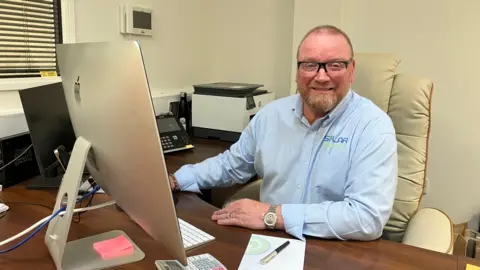 Terry Hayward sits at a desk in his office in front of a screen. He has a notebook and pen. He is wearing a light blue shirt with the company logo on it and glasses.