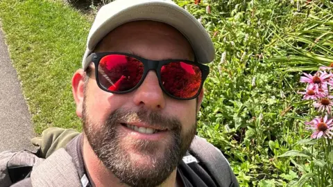 Image appears to be a selfie, James Desborough is wearing black rimmed sunglasses with red reflective lenses and is smiling at the camera. He is wearing a taupe baseball cap and appears to have a rucksack on. He has a dark beard and moustache. Behind him is grass and other green vegetation as well as some pink flowers.