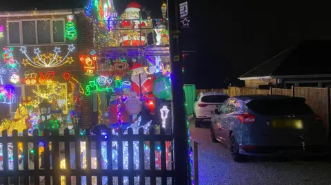 Bright Christmas lights are lit up on a house, with two cars parked next to the property