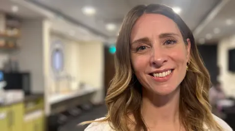 Harriet Heywood/BBC Ali Harris is indoors, standing in a brightly lit room with a modern and softly blurred background. She has long, wavy hair and is wearing a light-colored top along with earrings. Her face is turned toward the camera, and she’s smiling.