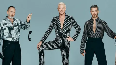 PA Media (l-r) Babydaddy, Jake Shears and Del Marquis, of the Scissor Sisters, posing in monochrome outfits in front of a pale blue background. Babydaddy holds an old-fashioned telephone receiver dangling by its cord from his right hand.