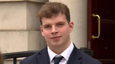 A young man looks into the camera. He wears a black suit with a tie and a white shirt. A big building is behind it.