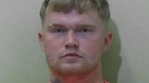 Northumbria Police Mugshot of Caris. He has faint blond stubble and short blond hair.