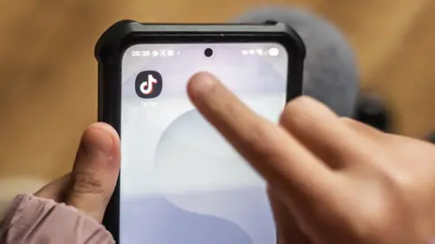 In the foreground is a child's finger reaching towards a TikTok logo on a phone screen. The phone is being held by their other hand. The background is out of focus but wooden flooring and dark coloured trousers can be made out.