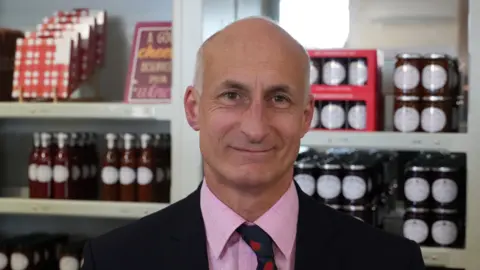 John Fairhall/BBC Chris Newnham is wearing a black suit jacket over a pink shirt and navy tie with red strawberries on it. Jam jars and glass bottles of sauce are stacked on shelves behind him.