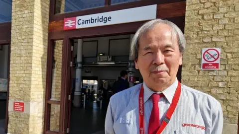 Greater Anglia Jeff Tan is standing outside of a brown bricked railway station which has a white sign above the door that reads "Cambridge". He has short grey hair and a moustache and is wearing a blue and white striped shirt, red tie and a red lanyard that says "Greater Anglia". 