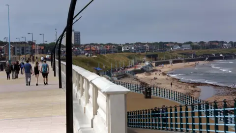 The promenade and coastline in Whitley Bay. People are strolling along the left-hand side of the picture, while the beach appears to the right.