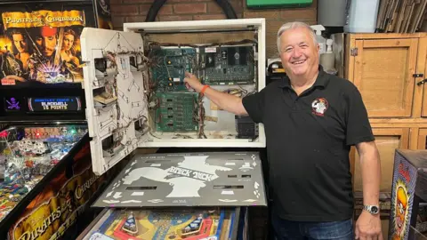 Jozef Hall/BBC Mark Squires with short white hair, wearing a grey polo shirt, smiling at the camera, holding up his right hand and pointing to wires and connections inside a pinball machine