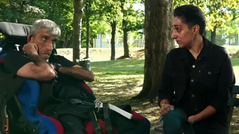 A picture of Amrik Reehal on the left, who has grey hair and is in a wheelchair and his sister Tajinder Blackman, who has black hair and is wearing a dark jacket is on the right. They are in a park with trees behind them. 