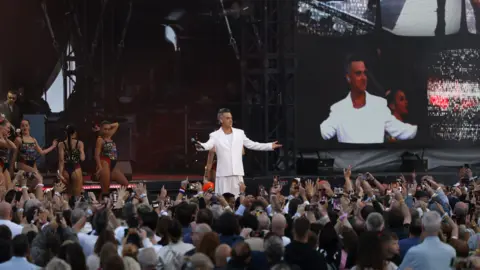 Getty Images Robbie Williams is dressed in an all-white suit on a stage in front of thousands. There are backing dancers dressed in sparkly costumes behind him.