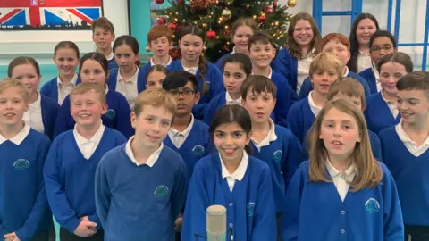 Winton Primary School A group of children in blue and white school uniform looking at the camera, with a microphone in front of them and a Christmas tree behind them.