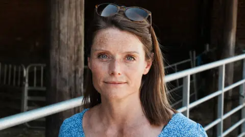John Fairhall/BBC Cath Crowther stands outside a farm pen with gates. She has shoulder length brown hair, some of which has been pushed back by sunglasses on top of her head. She wears a blue top with small white flowers printed all over it. 