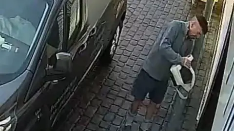 CCTV footage showing Maughan affixing a black box to a bollard using white tape. The bollard is outside a shop. Maughan is wearing shorts and has short hair which has been shaved completely around the back and sides of his head. He is standing behind a black van.