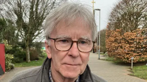 Shows a man with glasses and grey hair and wearing a brown leather jacket standing in front of trees
