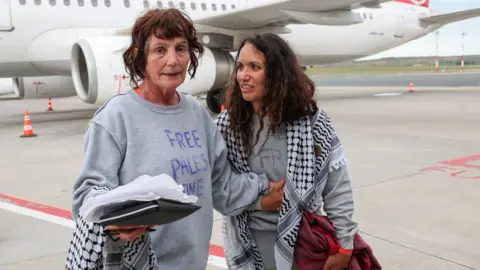 Reuters Two women stand on an airport tarmac beside a passenger plane, wearing grey sweatshirts with handwritten “Free Palestine” messages and black-and-white keffiyeh scarves. One looks toward the camera holding papers, while the other stands beside her carrying a red jacket.
