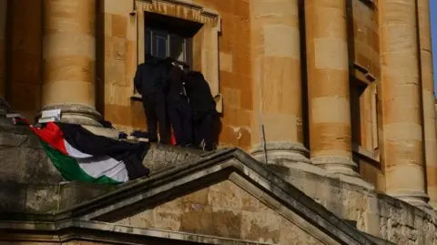 Four people all dressed in black on a ledge of the Radcliffe Camera are huddled around a window. A Palestinian flag hands from the ledge.