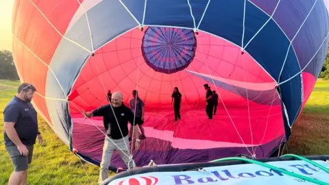 A picture of the inside of a partially inflated hot air balloon with five people inside and two men stood outside