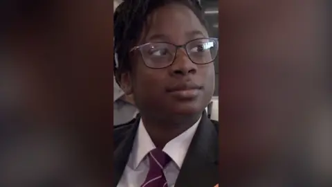 A young girl wearing school uniform, a white shirt and a black blazer. She has dark glasses.